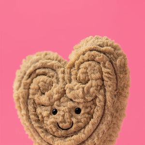 Jellycat light brown heart-shaped palmier plush toy with a smiling face on a pink background