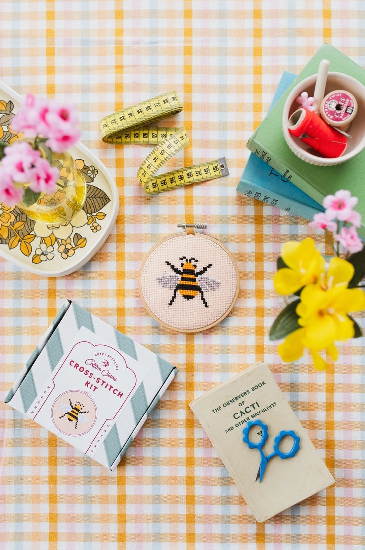 Cross-stitch kit with bee design contained in a circular embroidery hoop on a checkered tablecloth with flowers and books.