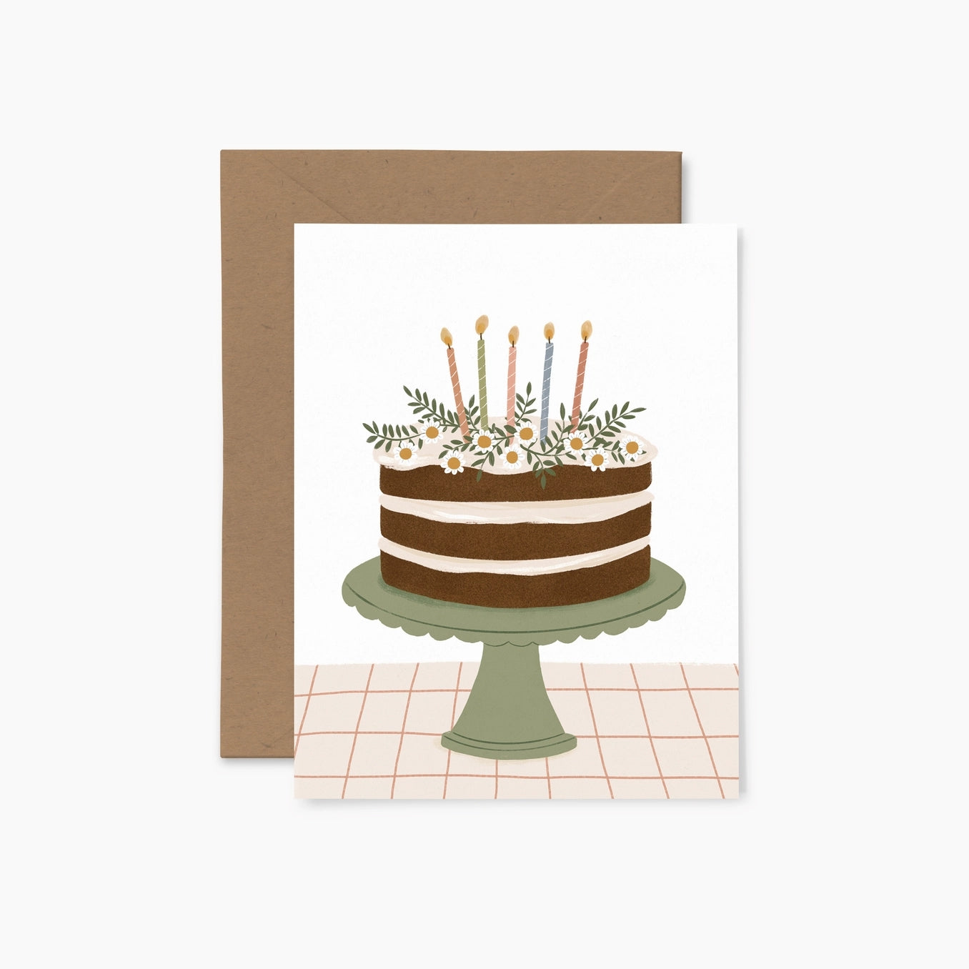 Greeting card featuring an illustration of a birthday cake sitting on top of a green cake stand. Five lit candles sit on top of the cake, along with some white and yellow flowers. The cake stand sits on top of a surface with a grid design against a white background. A brown envelope sits behind the card. 