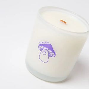 White candle with a wooden wick in a translucid glass with an image of a purple smiling mushroom and "Milkjar" written on top.