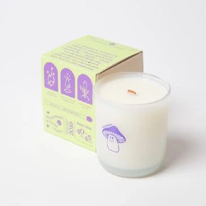 White candle in a translucid glass with showing an image of a purple smiling mushroom. The candle is placed beside a green packaging box with purple text and imagery. Both items are on a white background. 