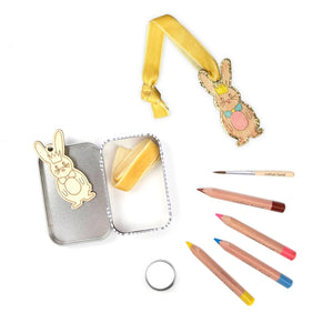 Open product packaging featuring a wooden bunny, yellow ribben, colouring pencils and a paintbrush, along with a bunny ornament on a white background.