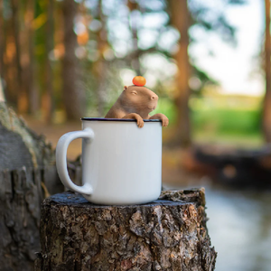 Image of a capybara tea infuser perched inside a white mug with a blurred nature background. 
