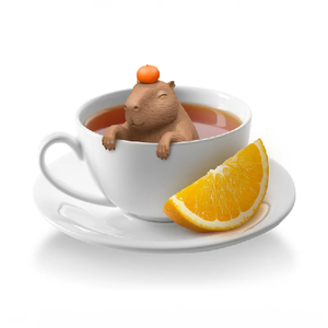 Capybara tea infuser perched inside a white mug filled with tea. The capybara is balancing an orange on it's head and the mug is sitting on a saucer, along with an orange slice.