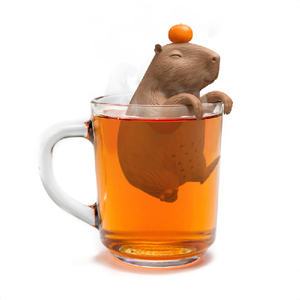 Capybara tea infuser perched inside a clear mug filled with tea on a white background. The capybara is balancing an orange on it's head.