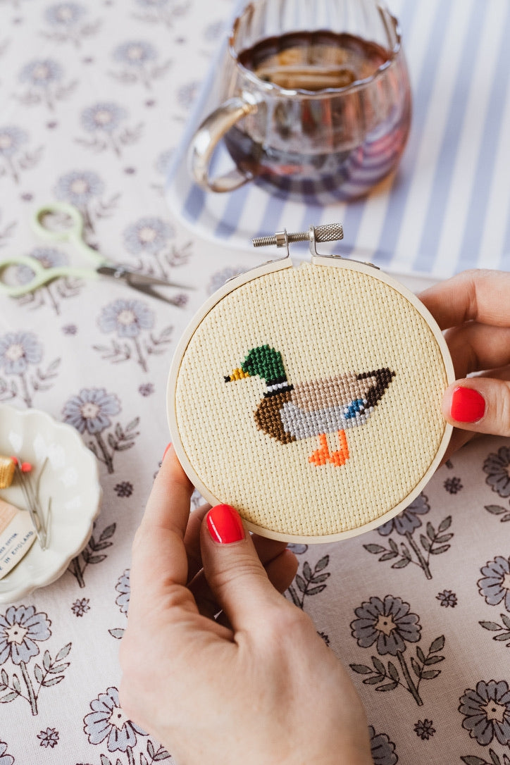 Hand holding a cross-stitched duck design in an embroidery hoop on a floral tablecloth with a teacup in the background.