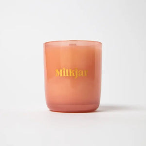 White candle in an orange glass with "Milkjar" written in yellow on a white background.