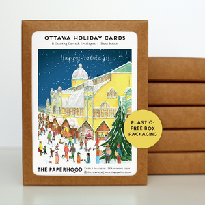 Boxed holiday cards featuring an illustration of Lansdowne Park in Ottawa with a Christmas theme, including people, trees, and a building with a snow-covered roof. In the background, more boxes are stacked on top of one another.