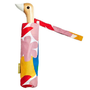 An umbrella with a colourful pink, red, yellow and blue abstract pattern with a duck head shaped handle on a white background. 