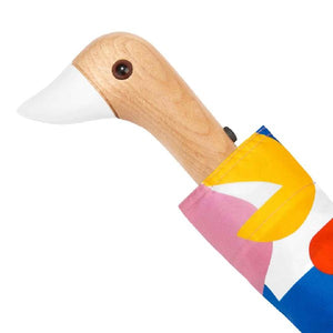 Closeup of an umbrella with a colourful pink, red, yellow and blue abstract pattern and a duck head shaped handle on a white background. 