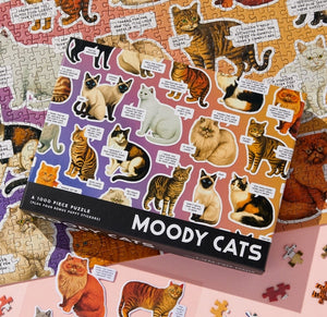 Packaging for a puzzle featuring colourful illustrations of cats with funny text on a pink and purple background. The packaging is placed on top of the completed puzzle on a tabletop.