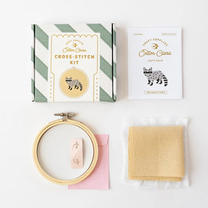 Cross-stitch kit with hoop, fabric, and an instructions manual on a white background.