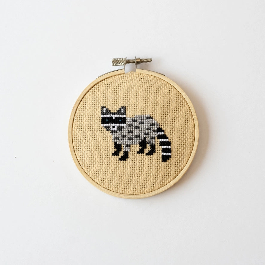 Cross-stitched raccoon design contained within a circular embroidery hoop against a white background.