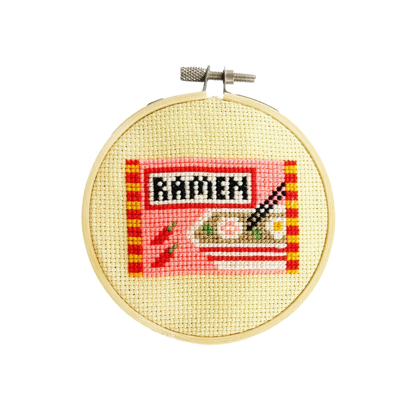 Cross-stitched design of a ramen pack in a wooden hoop on a white background.