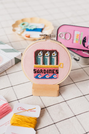 A cross-stitched design of sardines in a tin is framed in a wooden hoop. It sits on top of a surface with a grid design. 