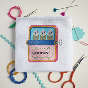 Cross-stitched design of a tin of sardines with the text 'sardines', surrounded by crafting materials on a light background.
