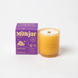 Canadian company Milkjar candle and its box packaging. The white candle is in an yellow glass with "Milkjar" written in gold. The other side of the candle has a cute smiling cloud with legs doodle in gold. The packaging is a small purple box with yellow text and graphics. 