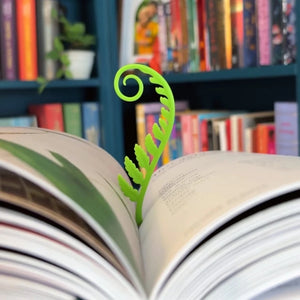 Green fern-shaped bookmark in an open book with a bookshelf background.